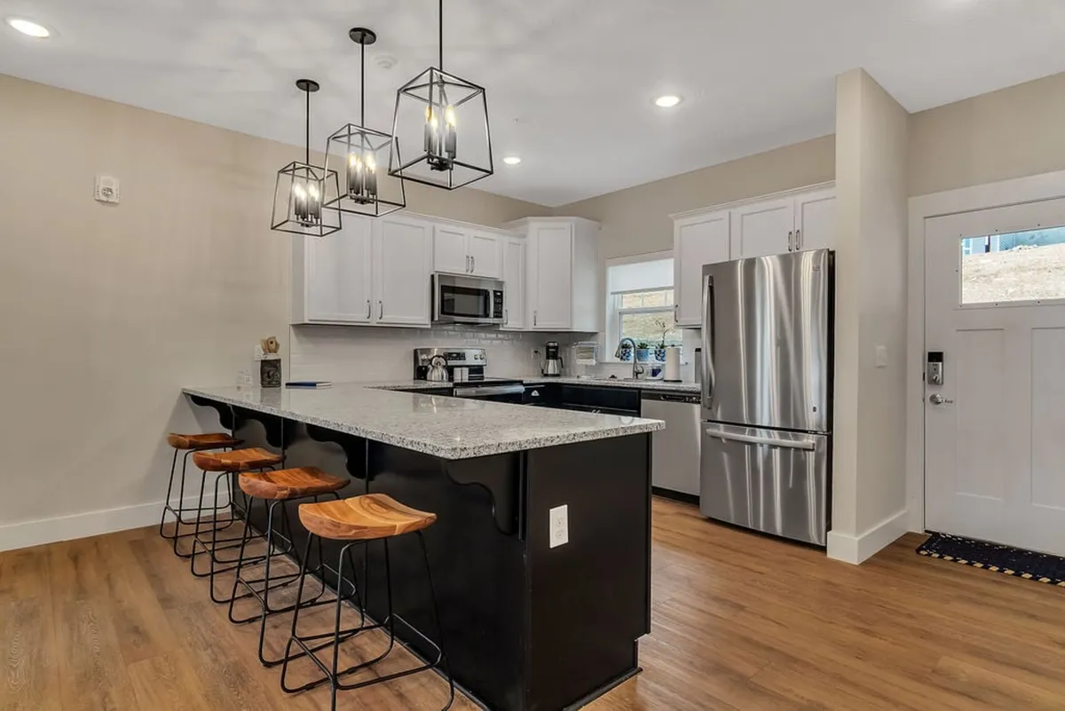 A modern, fully-equipped kitchen with a prominent island bar and stools, gleaming stainless steel appliances, striking granite countertops, and stylish hanging light fixtures. The layout is open and welcoming, ideal for cooking and casual dining.