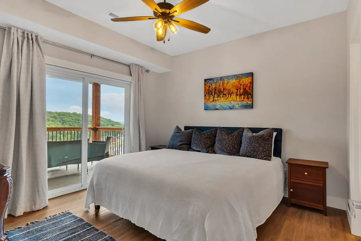 A serene bedroom with a large bed, colorful artwork, and a door that opens onto a balcony with a view. The space blends restful interiors with direct outdoor access, suggesting the allure of both comfort and scenery.