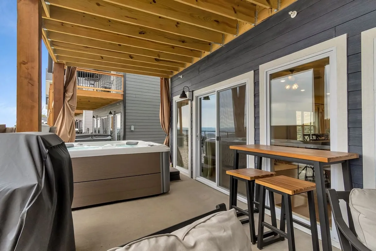 A serene exterior shot of a private hot tub set on a wooden deck, with a bar table and sliding glass doors that connect the patio to the interior of the home. The surrounding landscape enhances the sense of privacy and tranquility, while the space is clearly designed for entertaining and relaxation.