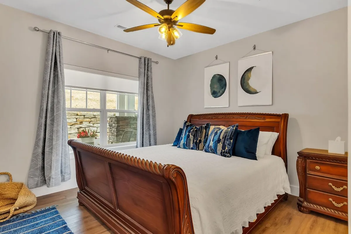 A serene bedroom with a wooden bed frame, decorative pillows, artwork on the wall, and a window with curtains letting in natural light. The ambiance is calm and restful, suggesting a retreat ideal for relaxation.