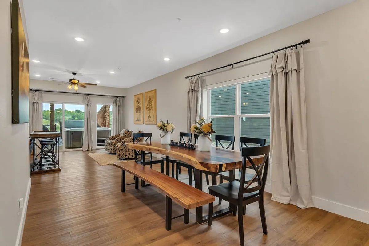 The dining area centers around a long, striking wood slab dining table with decorative vases filled with flowers. Large windows flood the room with natural light, and ample seating is perfect for communal meals and shared stories.