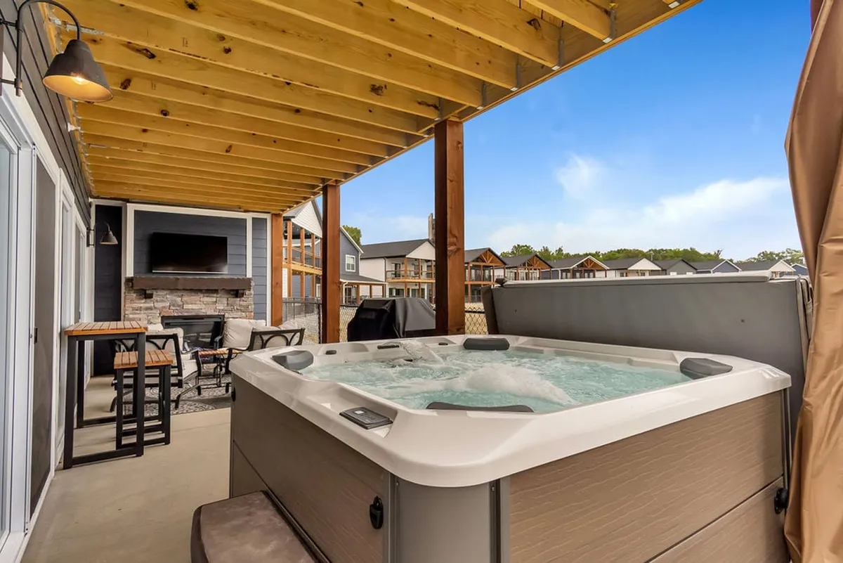 This image showcases a cozy covered patio with a hot tub, outdoor seating, and a fireplace framed by wooden beams. The space is inviting, with plush chairs arranged for conversation and relaxation, and the hot tub positioned to take in the outdoor ambiance—possibly with a view of the sunset or lake beyond.