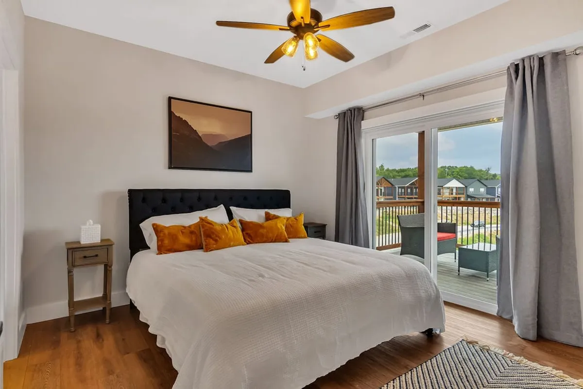 A comfortable bedroom featuring a bed with colorful pillows, ceiling fan, sliding glass door to a balcony, and wall art. The sliding door reveals balcony access and the promise of scenic views, aligning with the upstairs king suites' unique features.